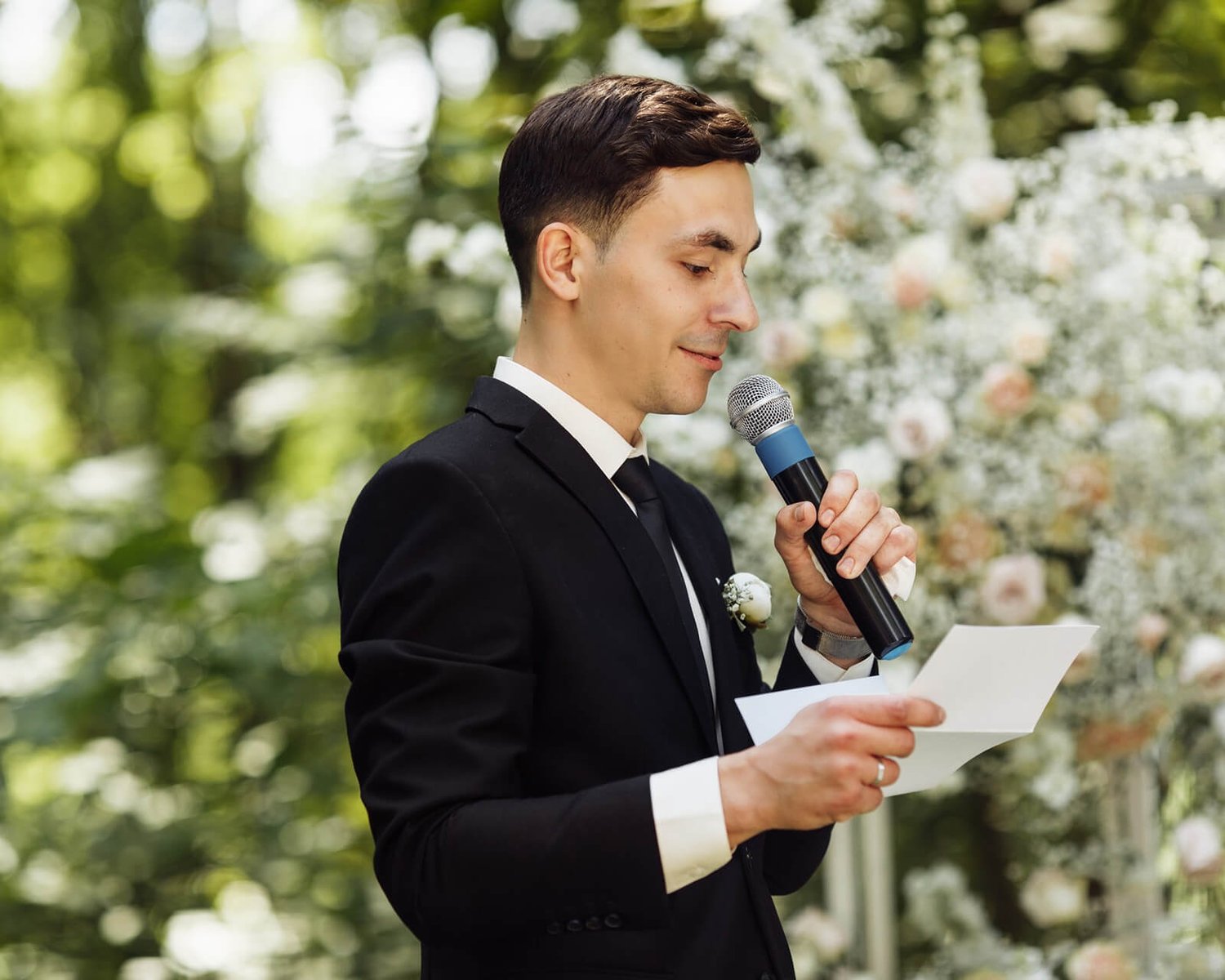 Groom delivering a wedding speech outdoors, holding a microphone and a note card, with a floral backdrop.