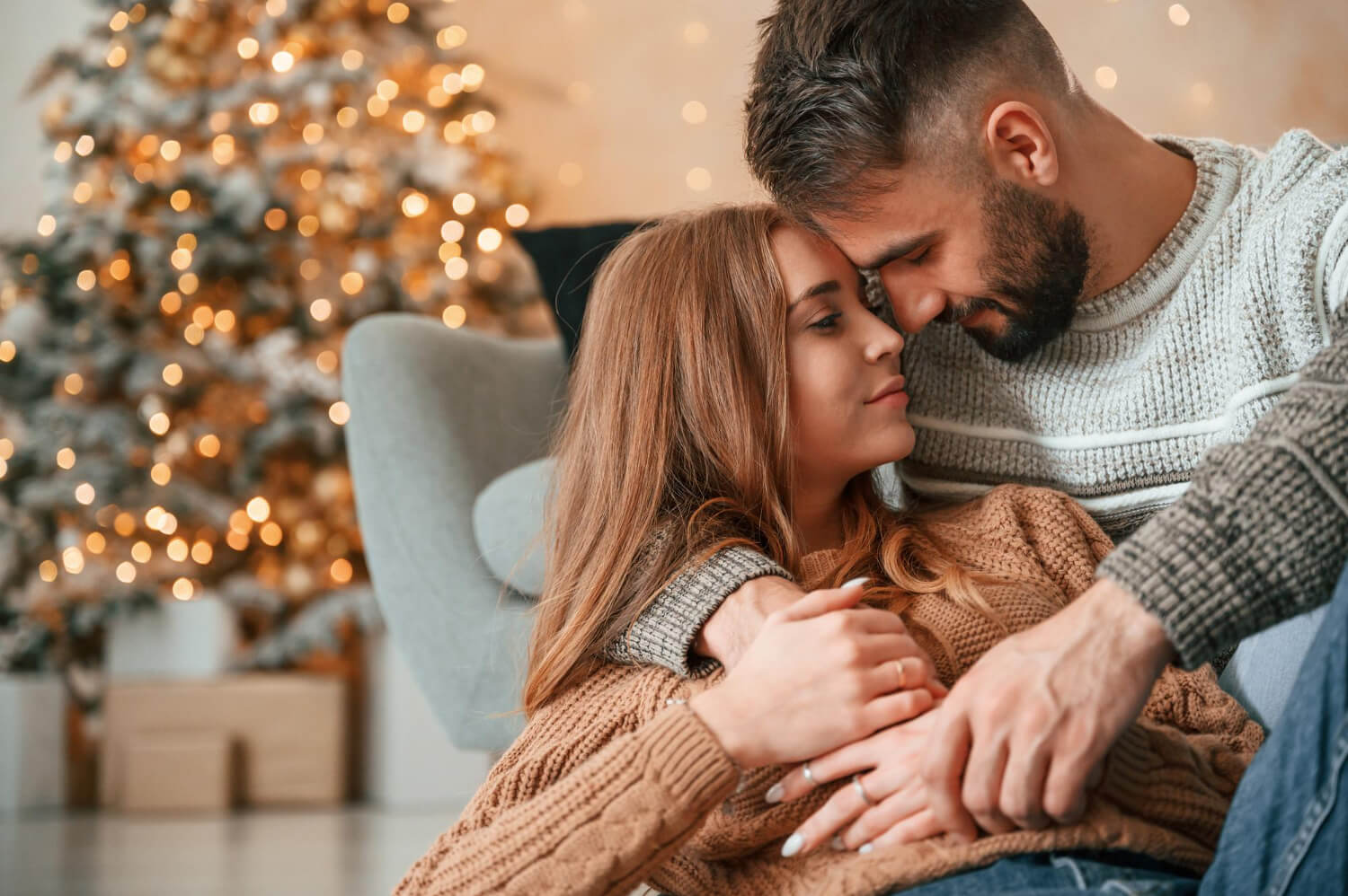 a couple romantically sitting together, the man is holding the woman in his arms, there is a christmas tree in the background