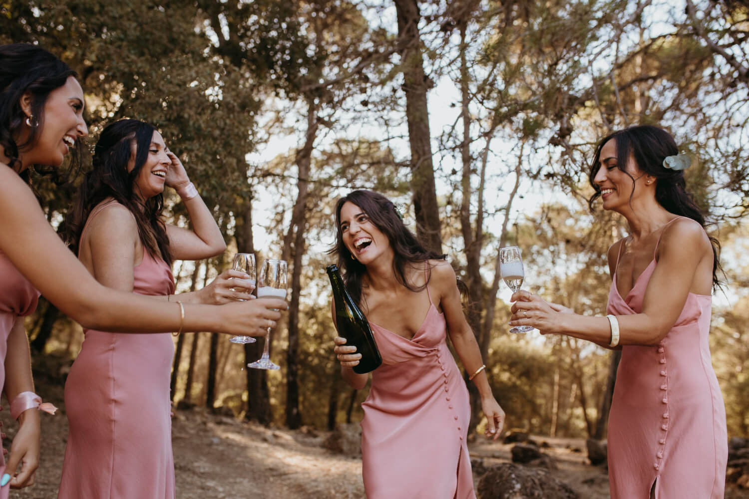 A group of women standing outside in a forest area. They are all dressed in pink dresses. One woman is holding a champagne bottle and the others holding glasses. They seem drunk and are all laughing.