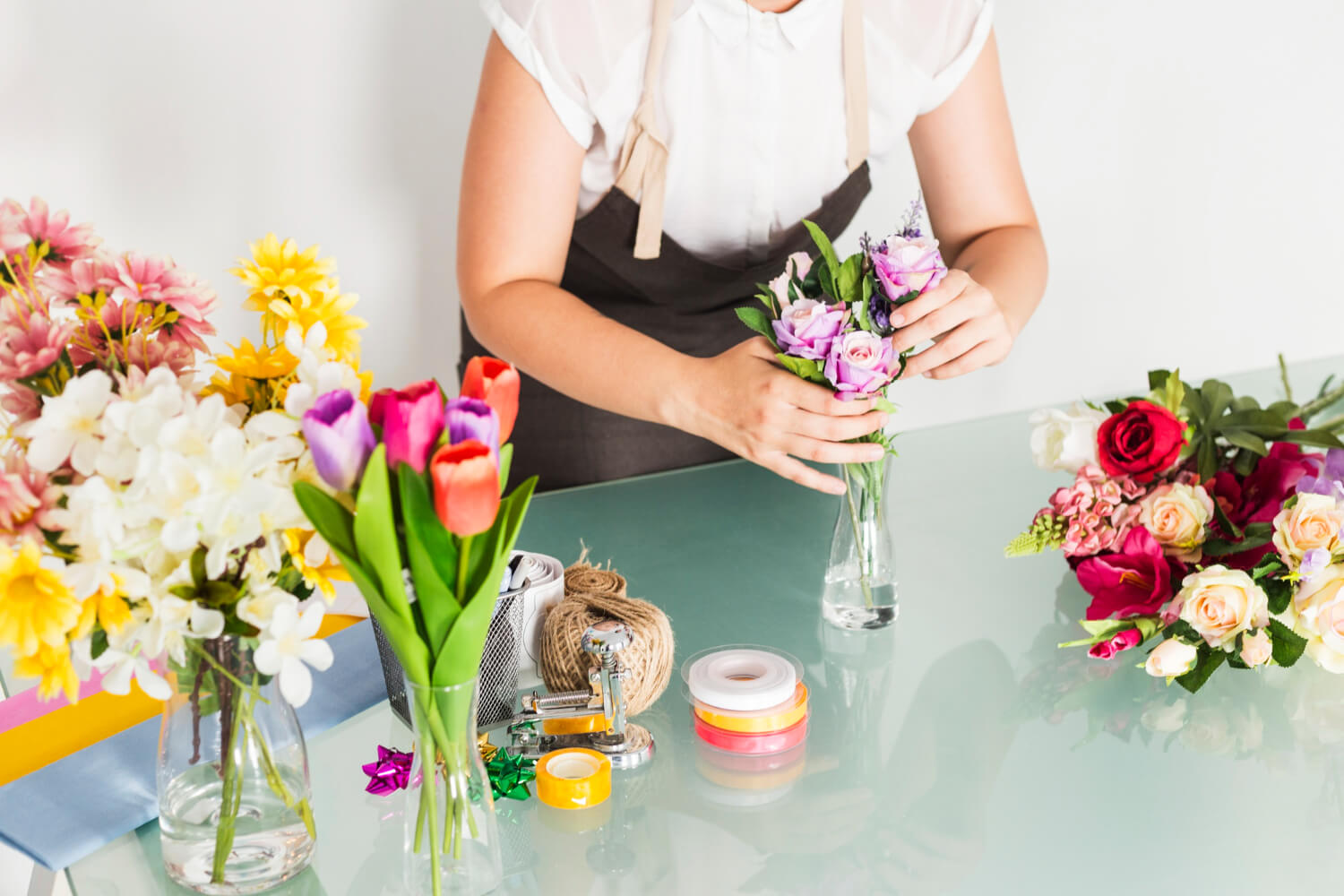 woman standing in front of a desk with craft tools and flowers is creating a flower bouquet