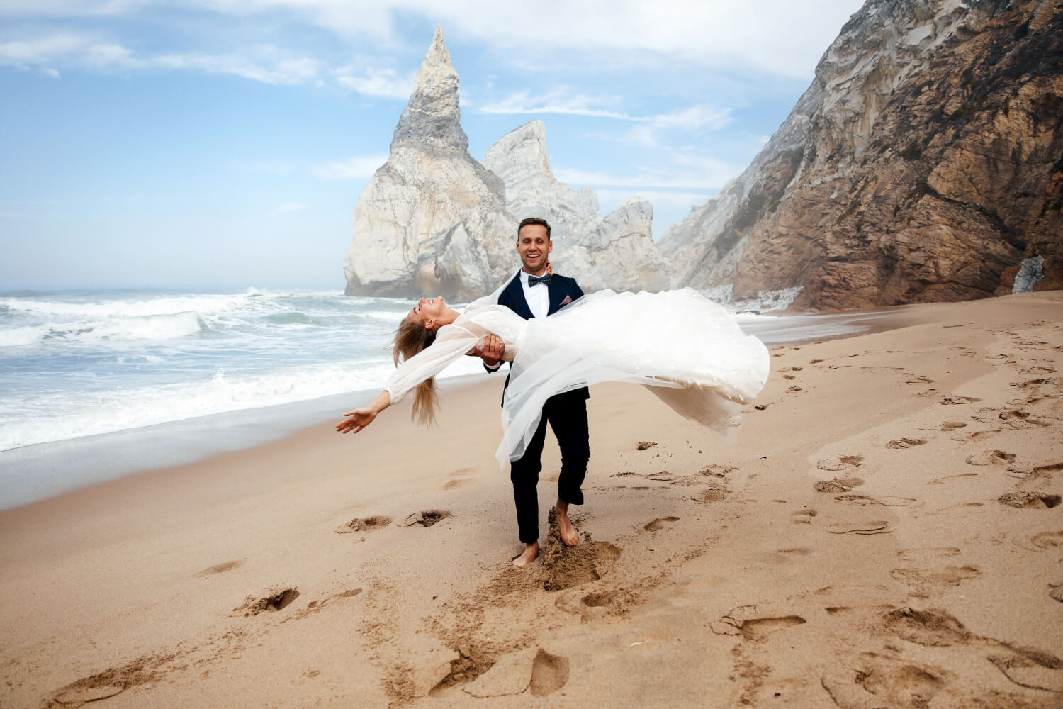 the groom carries the bride in his arms as they walk along the beach