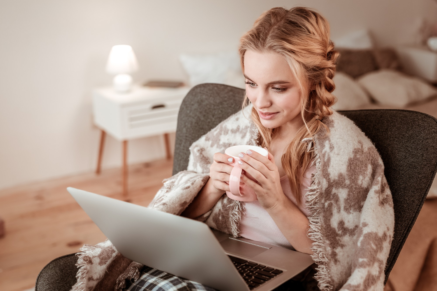 woman sitting on a chair, drinking coffee, and looking at a laptop screen