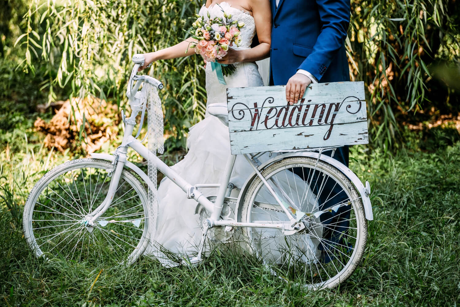 bride and groom standing next to a white bicycle with a wooden wedding sign