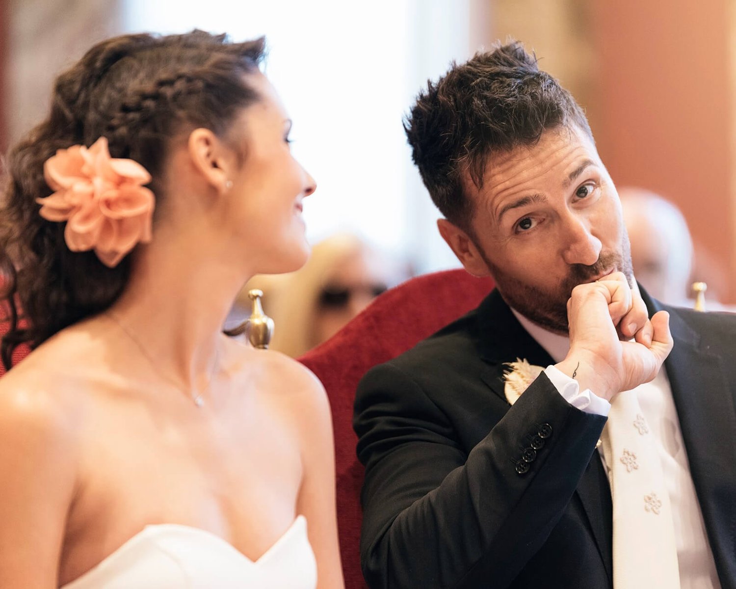 Wedding couple listening attentively, with the groom holding his chin and the bride smiling beside him.