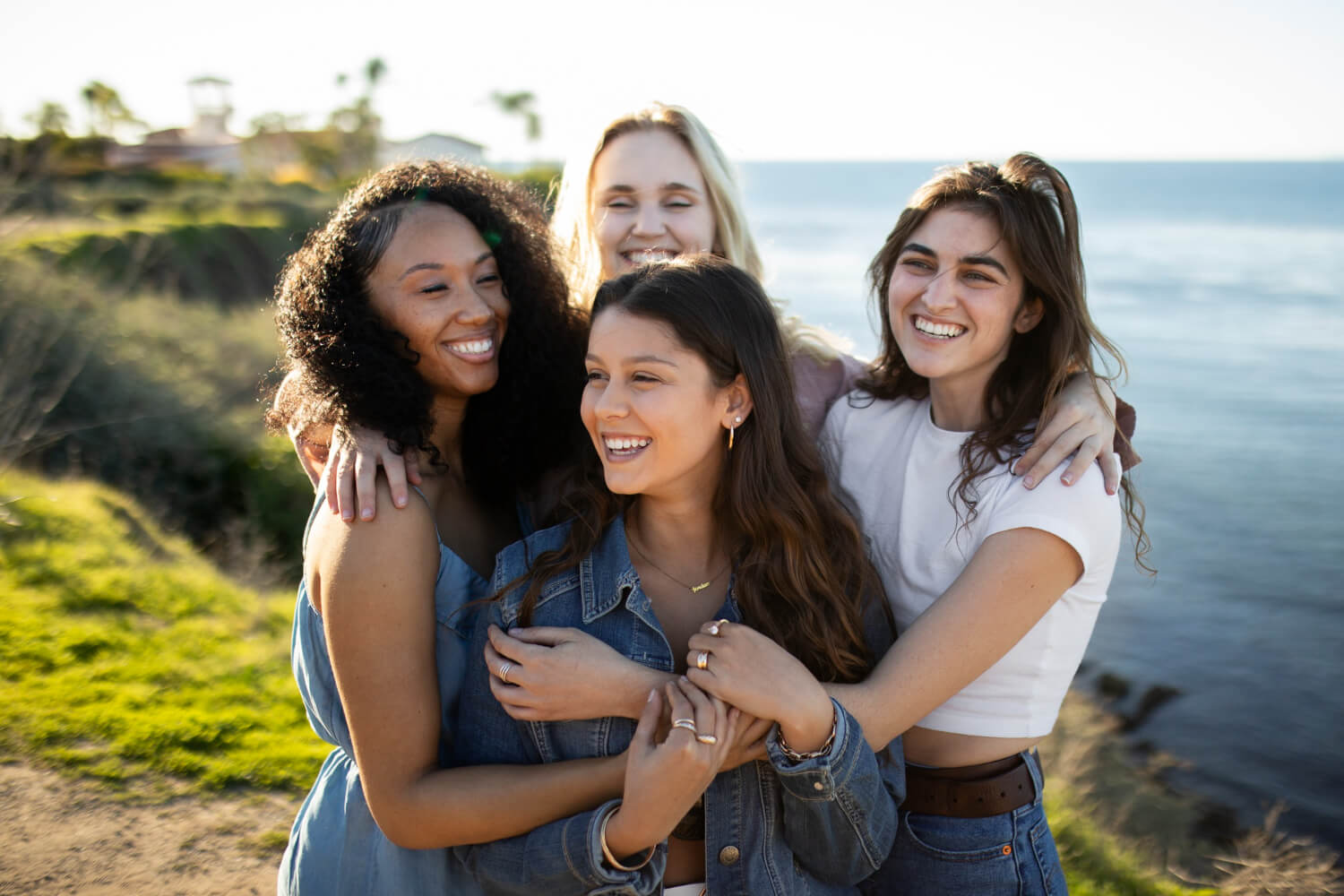 four women standing together outside near the ocean hugging each other