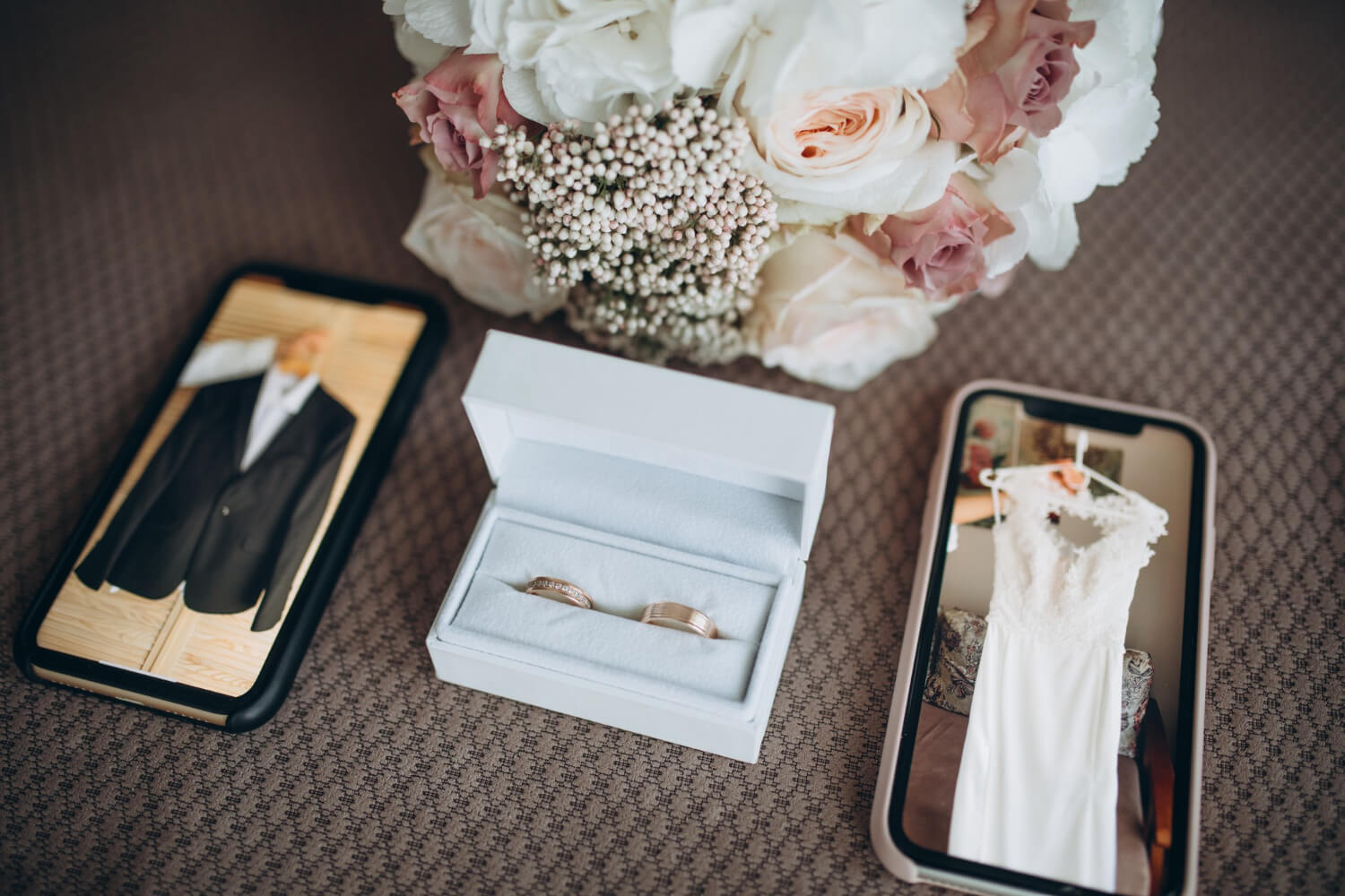 a box with two wedding rings between two phones that show the grooms suit and the brides dress on their screens