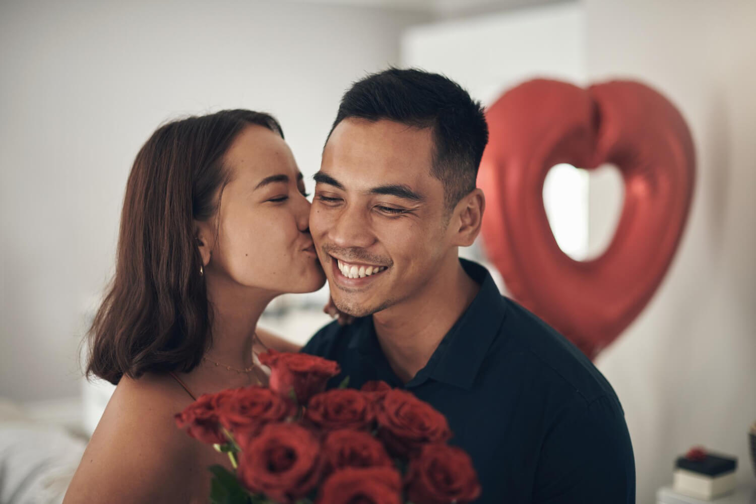 a man and a woman, both are happy, she is holding a bouquet of red roses, she is kissing him on the cheek